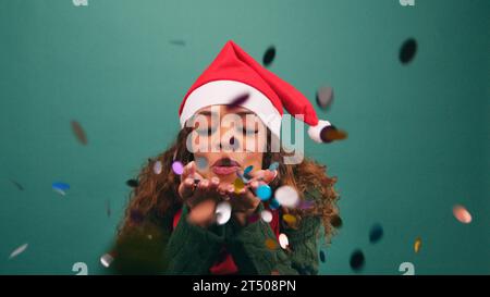 Belle jeune femme souffle des confettis arc-en-ciel à la caméra, studio de Noël Banque D'Images