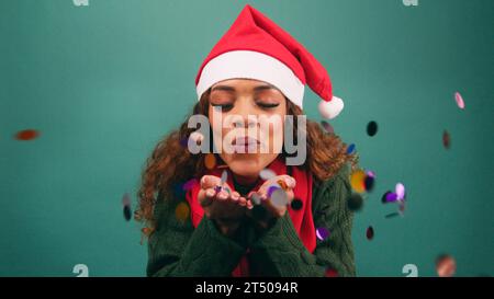 Belle jeune femme souffle des confettis arc-en-ciel à la caméra, studio de Noël Banque D'Images