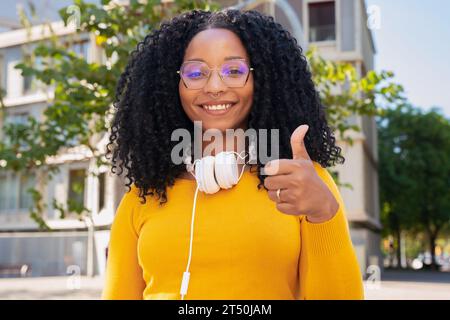 Portrait Jeune femme afro-américaine pointant le pouce vers le haut regardant souriant à la caméra à l'extérieur. Banque D'Images