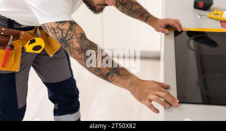 l'homme met la plaque de cuisson dans la cuisine. Installation d'une table de cuisson à induction au-dessus du four. Travailler conformément aux normes et standards techniques. Service de réparation rapide et Banque D'Images