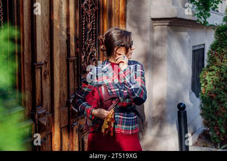 Portrait d'une femme élégante portant une pince à cheveux tenant un sac à main rouge portant un blazer à carreaux, accessoires sur la rue de la ville d'automne. Automne à la mode vieux m Banque D'Images