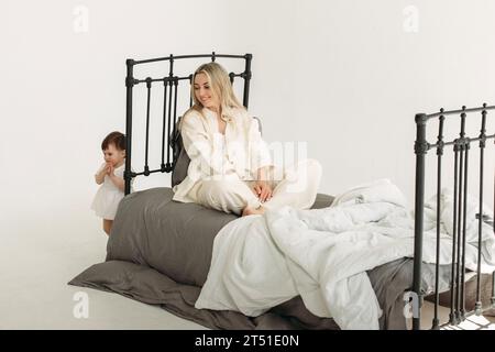 Une mère heureuse s'assoit sur le lit et regarde sa fille d'un an courir. Concept de famille. Banque D'Images