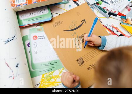 Paris, France - 25 octobre 2023 : d'en haut, un enfant curieux dessine les yeux sur un logo Amazon à l'aide de crayons. Sur sa table créative, la scène suggère un intérêt grandissant pour le graphisme. Banque D'Images