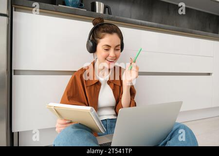 La femme étudie, fait ses devoirs à la maison, se connecte à la leçon en ligne, s'assoit sur le sol avec un ordinateur portable, écrit dans un ordinateur portable, travaille dans un casque Banque D'Images