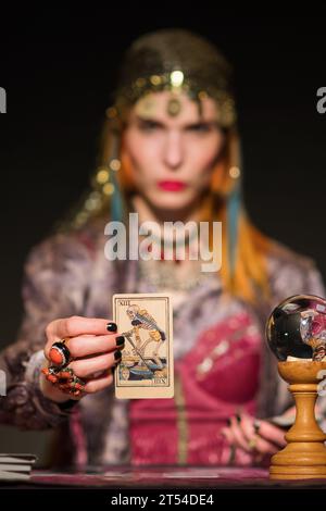 Brouillée diseuse de fortune féminine dans le maquillage et tenue élégante regardant la caméra tout en étant assis à table avec boule de verre et montrant la carte de décès contre Banque D'Images