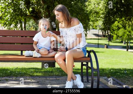 Mère et fille assis sur un banc dans le parc de la ville d'été et soufflant des bulles de savon . Enfance, loisirs et concept de personnes - repos familial heureux an Banque D'Images
