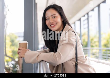 Image vue de côté d'une belle et attrayante femme d'affaires asiatique millénaire boit du café sur le Skywalk, prenant une pause café rapide avant d'aller Banque D'Images