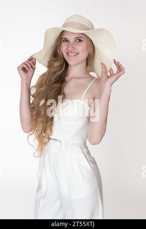 Expression faciale blondie Femme mordant la lèvre, les bras levés et tenant le bord d'un chapeau de paille souple avec les deux mains. Portrait femme ludique bien habillée Banque D'Images