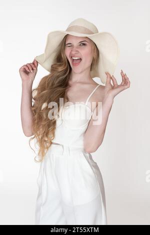 Femme sourit la bouche ouverte, un œil fermé et clignant à la caméra. Expression faciale d'une femme levant les bras et tenant le bord d'un chapeau de paille souple Banque D'Images