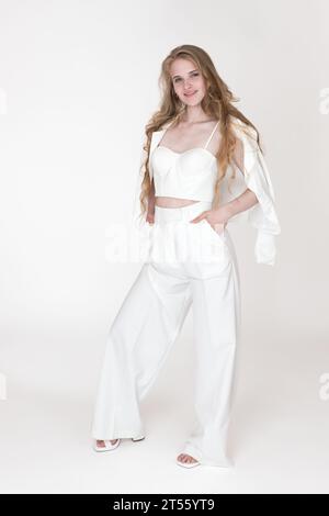Femme souriante élégamment habillée en veste de costume blanc, sculptant haut de corset coupe, pantalon, frondes debout sur fond blanc Banque D'Images