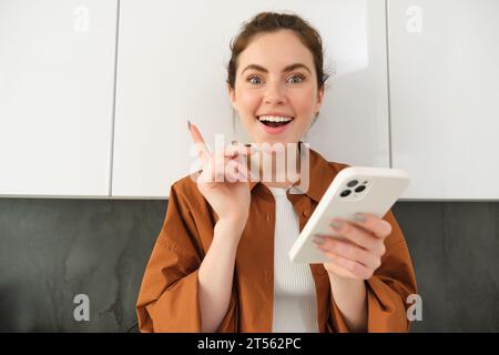 Portrait de jeune femme excitée a une idée, tenant le smartphone et pointant le doigt vers le haut, debout dans la cuisine à la maison Banque D'Images