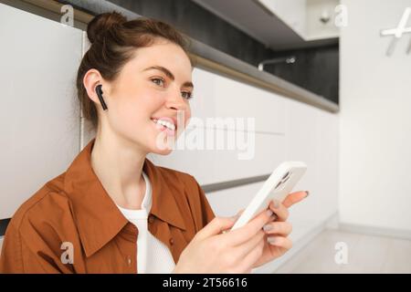 Portrait de jeune femme moderne dans un casque sans fil, assis sur le sol de la cuisine, en utilisant un smartphone, en faisant une playlist avec des chansons préférées, écoute Banque D'Images