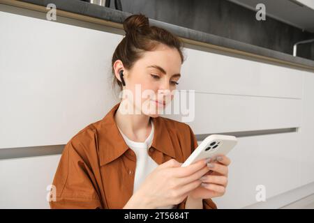 Portrait de jeune femme moderne dans un casque sans fil, assis sur le sol de la cuisine, en utilisant un smartphone, en faisant une playlist avec des chansons préférées, écoute Banque D'Images