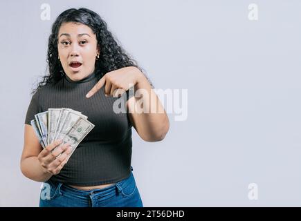 Jeune femme étonné tenant et pointant des billets de banque en dollars isolés. Jeune fille latine surprise détenant de l'argent liquide isolé Banque D'Images