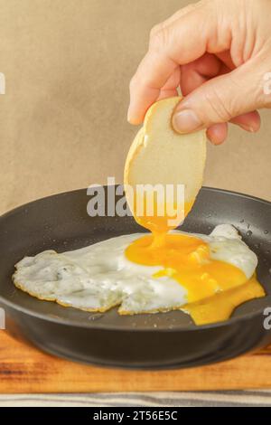 Femme main trempant du pain sur le jaune d'un œuf frit dans l'huile d'olive avec une poêle sur une planche de bois Banque D'Images