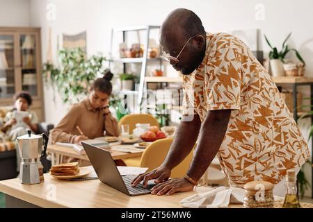 Homme noir sérieux dans les lunettes vérifiant les e-mails sur ordinateur portable lorsqu'il travaille à domicile le week-end Banque D'Images