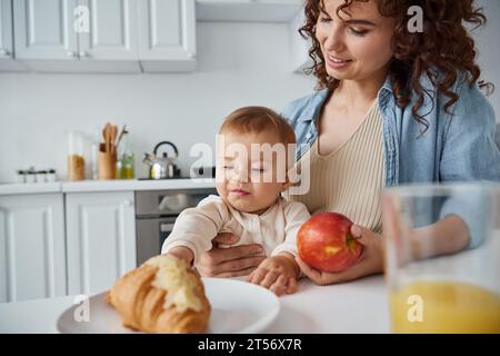 femme souriante avec pomme mûre près de la petite fille atteignant délicieux croissant, petit déjeuner dans la cuisine Banque D'Images