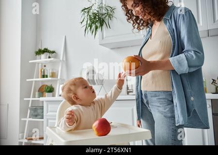 mignon enfant dans la chaise bébé atteignant orange mûr à la main de mère souriante dans la cuisine, repas Banque D'Images