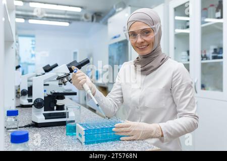 Étudiante musulmane en chimie en hijab assise à une table dans un laboratoire de recherche et étudiant des substances liquides, tenant un bécher avec une substance et souriant à la caméra. Banque D'Images