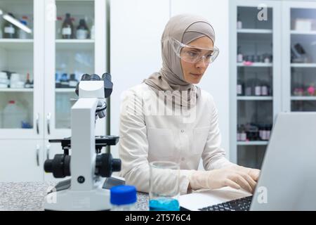 Jeune femme arabe en hijab, médecin assise à une table et travaillant sur un ordinateur portable dans un laboratoire. Banque D'Images