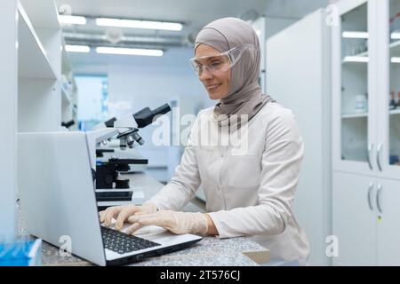 Jeune scientifique arabe, pharmacienne en hijab assise à table avec microscope, liquides en bouteille et travaillant sur ordinateur portable. Banque D'Images