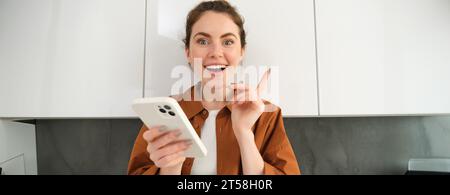 Portrait de jeune femme excitée a une idée, tenant le smartphone et pointant le doigt vers le haut, debout dans la cuisine à la maison Banque D'Images