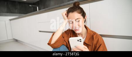 Portrait de jeune femme moderne dans un casque sans fil, assis sur le sol de la cuisine, en utilisant un smartphone, en faisant une playlist avec des chansons préférées, écoute Banque D'Images