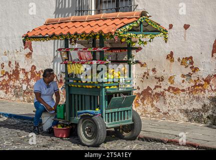 Guatemala, la Antigua - 20 juillet 2023 : un vendeur masculin de fruits de rue est assis avec sa poussette verte le long du côté de la rue. Mur beige avec des taches rouillées comme dos Banque D'Images