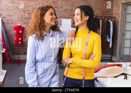 Deux créatrices de mode biraciales heureuses debout avec du ruban à mesurer et riant en studio. Mode, design, créativité, couture, vêtements et Banque D'Images