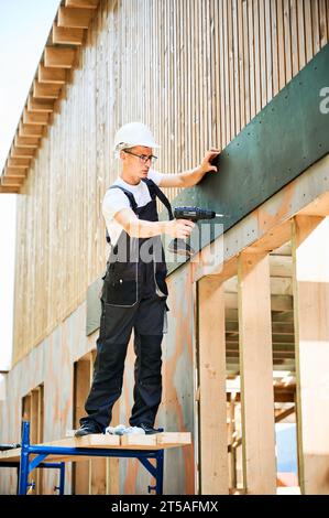 Carpenter construisant une maison en bois encadrée. Homme ouvrier en lunettes habillage façade de maison avec panneaux de particules de ciment, les fixant avec des tournevis. Concept de construction écologique moderne. Banque D'Images