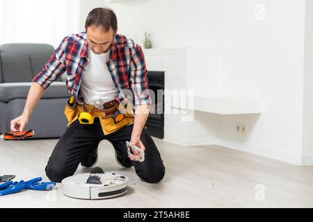 Un jeune homme de race blanche effectue la maintenance d'un robot-aspirateur blanc. Aspirateur moderne sans fil autonome. Nettoyant robot de réparation pour homme à la maison Banque D'Images