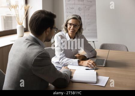 Heureuse femme d'affaires mature parlant à un collègue masculin plus jeune à l'ordinateur portable Banque D'Images