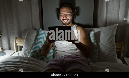 Jeune homme arabe utilisant le touchpad assis sur le lit portant des écouteurs dans la chambre Banque D'Images