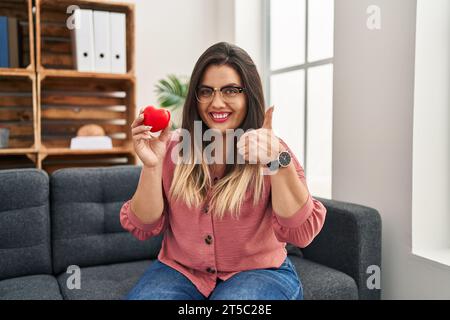 Jeune femme hispanique travaillant sur la thérapie de couple au bureau de consultation souriant heureux et positif, pouce vers le haut faisant excellent et signe d'approbation Banque D'Images