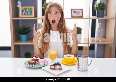 Jeune femme caucasienne mangeant des pâtisseries t pour le petit déjeuner étonné et surpris regardant vers le haut et pointant avec les doigts et les bras levés. Banque D'Images