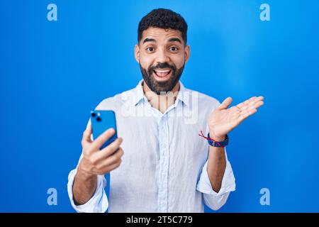 Homme hispanique avec barbe à l'aide de smartphone tapant message pointant de côté avec les mains ouvertes paumes montrant l'espace de copie, présentant la publicité souriante exci Banque D'Images