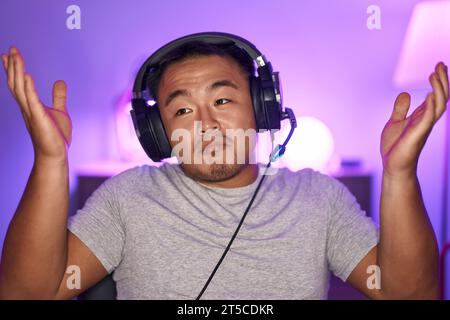 Jeune homme chinois jouant à des jeux vidéo portant des écouteurs criant et criant fort sur le côté avec la main sur la bouche. concept de communication. Banque D'Images