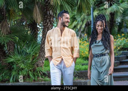 Un jeune couple à la mode profite d'un moment de compagnie détendue tout en marchant dans un cadre de jardin animé, leurs tenues estivales élégantes complètent Banque D'Images