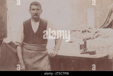 Photographie du début du 20e siècle d'un charpentier ou menuisier debout près de son établi. Le menuisier porte une toile ou un tablier hessien sur sa chemise et son gilet. Des outils sont sur l'établi, y compris un avion et un maillet, ainsi que quelques petits morceaux de bois et une pile de copeaux de bois. Un grand sac est suspendu à gauche de l'homme et d'autres sacs sont empilés à l'arrière du banc Banque D'Images