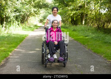 Femme indienne asiatique poussant sa mère âgée dans un fauteuil roulant à l'extérieur en été, Royaume-Uni. Peut également représenter un soignant, soins dans la communauté Banque D'Images