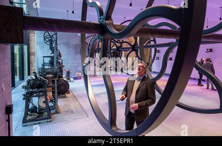 PRODUCTION - 03 novembre 2023, Saxe, Plauen : Martin Salesch, directeur du musée, se tient à côté d'une poinçonneuse (l) au milieu de l'exposition dans le nouveau musée de la Maison Weisbach à Plauen. En tant que «usine de fils», le nouveau complexe muséal dans l'un des plus anciens et premiers bâtiments de manufacture de Saxe ouvre ses portes aux visiteurs le 12 novembre. Les tissus textiles, la broderie et surtout le plus grand succès d'exportation de la ville, 'Plauen Lace', seront au centre de l'exposition. Des installations lumineuses avec des fils, de la dentelle et de la broderie caractérisent l’exposition. Machine à broder historique Banque D'Images