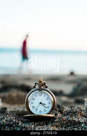 Montre de poche en or se trouve sur une plage de sable avec une personne marchant sur la plage en arrière-plan. Avec espace de copie pour le texte Banque D'Images