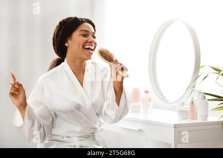 Joyeuse femme afro-américaine chantant avec brosse à cheveux tout en étant assise à la coiffeuse Banque D'Images