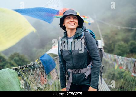 Jeune femme routard souriante traversant le canyon sur le pont suspendu avec des drapeaux de prière tibétains multicolores. Route d'escalade de pic de Mera trek Lukla Saga Banque D'Images