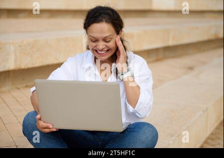 Heureuse femme aux cheveux bouclés souriant tout en travaillant sur un ordinateur portable à l'extérieur assis sur des marches. Personnes. Style de vie. Commerce en ligne. Banque D'Images