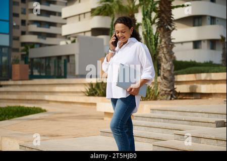 Femme multi ethnique confiante en chemise décontractée blanche, tenant un ordinateur portable, parlant sur un téléphone mobile intelligent contre fond de paysage urbain. Personnes. Communication. Banque D'Images