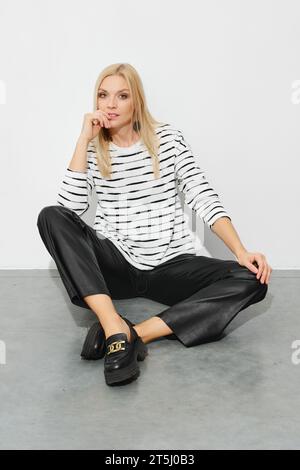 Femme blonde tendance dans un blazer rayé et pantalon en cuir large se trouve sur le sol Banque D'Images