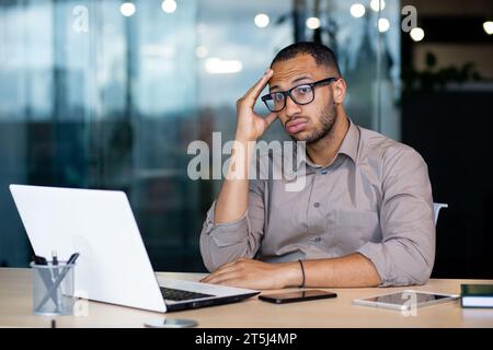 Frustré et triste homme au lieu de travail lisant le message en ligne de l'ordinateur portable, homme d'affaires assis à l'intérieur du bureau au lieu de travail, désespéré malheureux, a eu de mauvaises nouvelles, faillite, licenciement. Banque D'Images