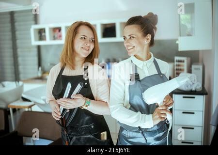 sourire deux femmes de 40 ans coiffeurs dans des tabliers dans un salon de beauté moderne avec lisseur et sèche-cheveux. Banque D'Images