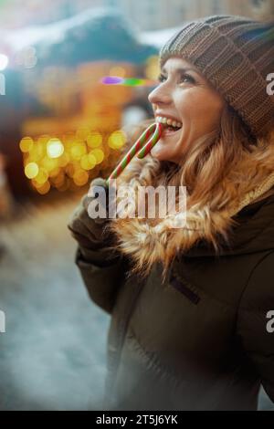 heureuse jeune femme en manteau vert et chapeau brun à la foire d'hiver dans la ville avec canne à sucre. Banque D'Images